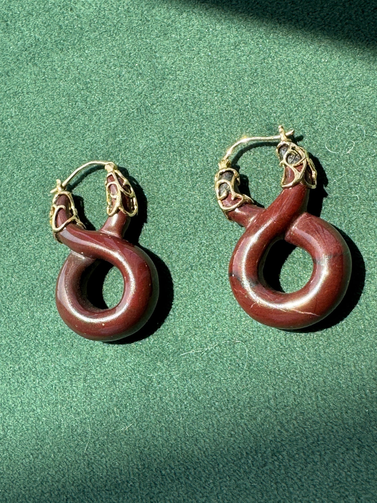 A pair of hoop earrings with dark reddish-brown curved stone pendants and gold-toned metal hoops, displayed on a textured green surface.