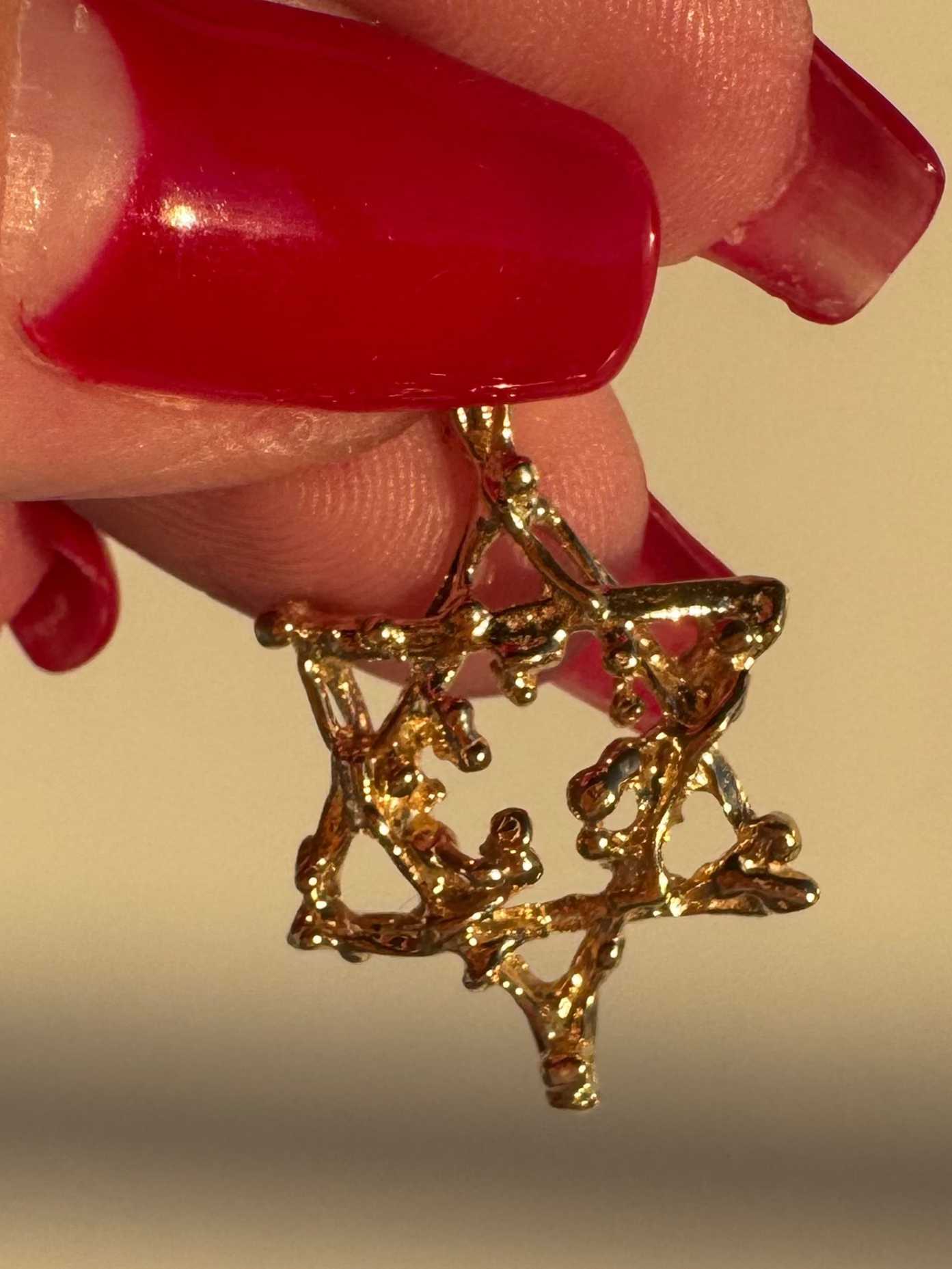 Gold earrings held by a hand with red nail polish against a beige background