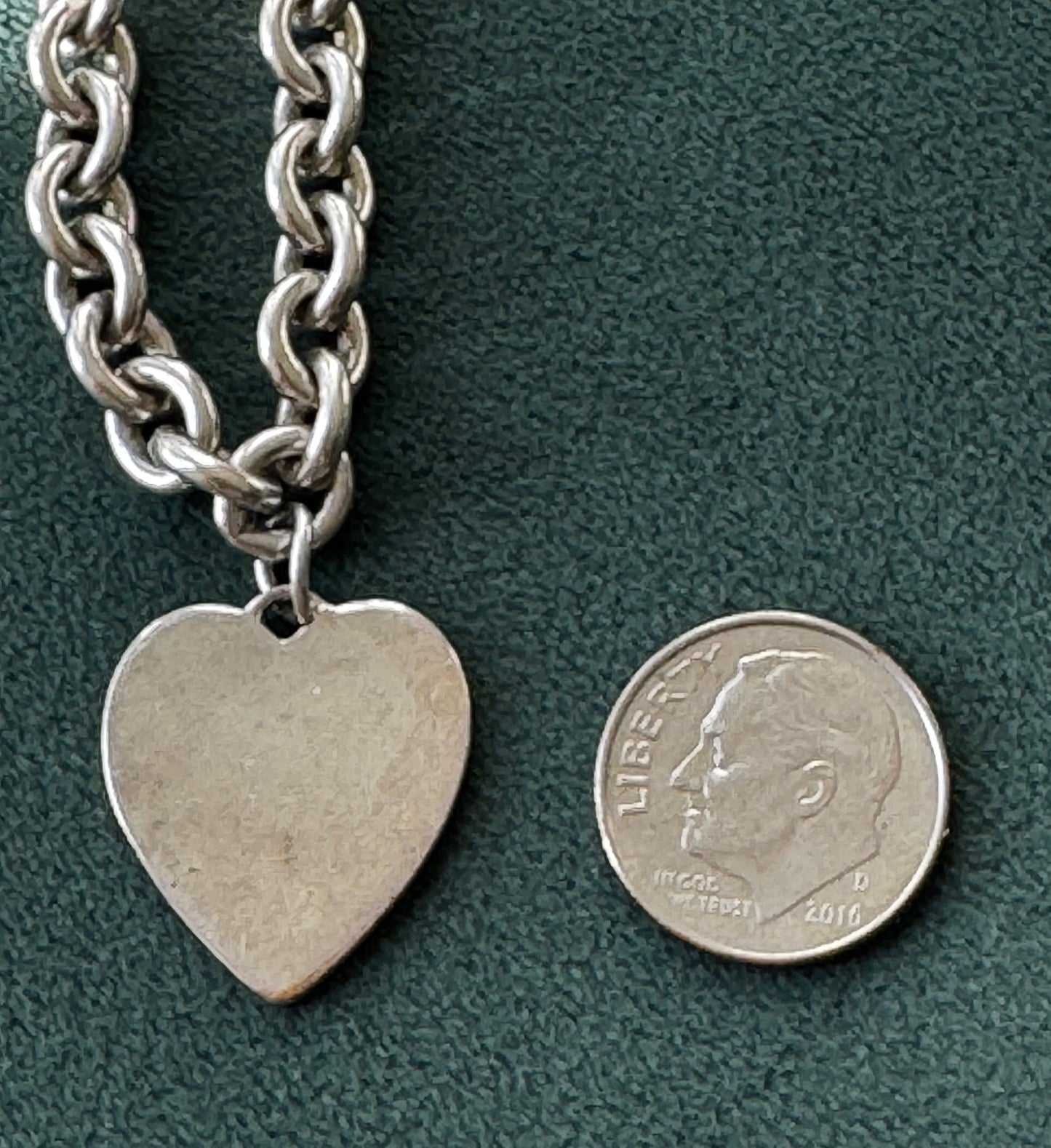 Silver chain bracelet with a heart-shaped tag next to a coin on a green surface