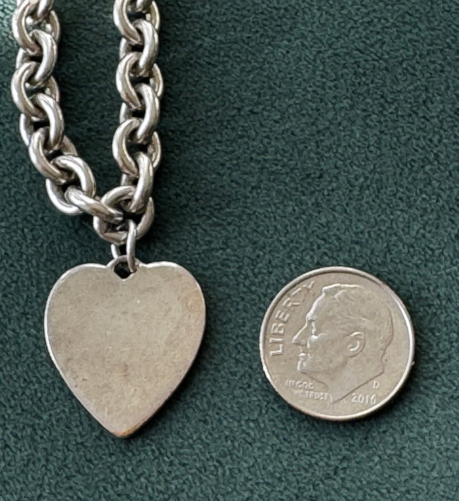 Silver chain bracelet with a heart-shaped tag next to a coin on a green surface