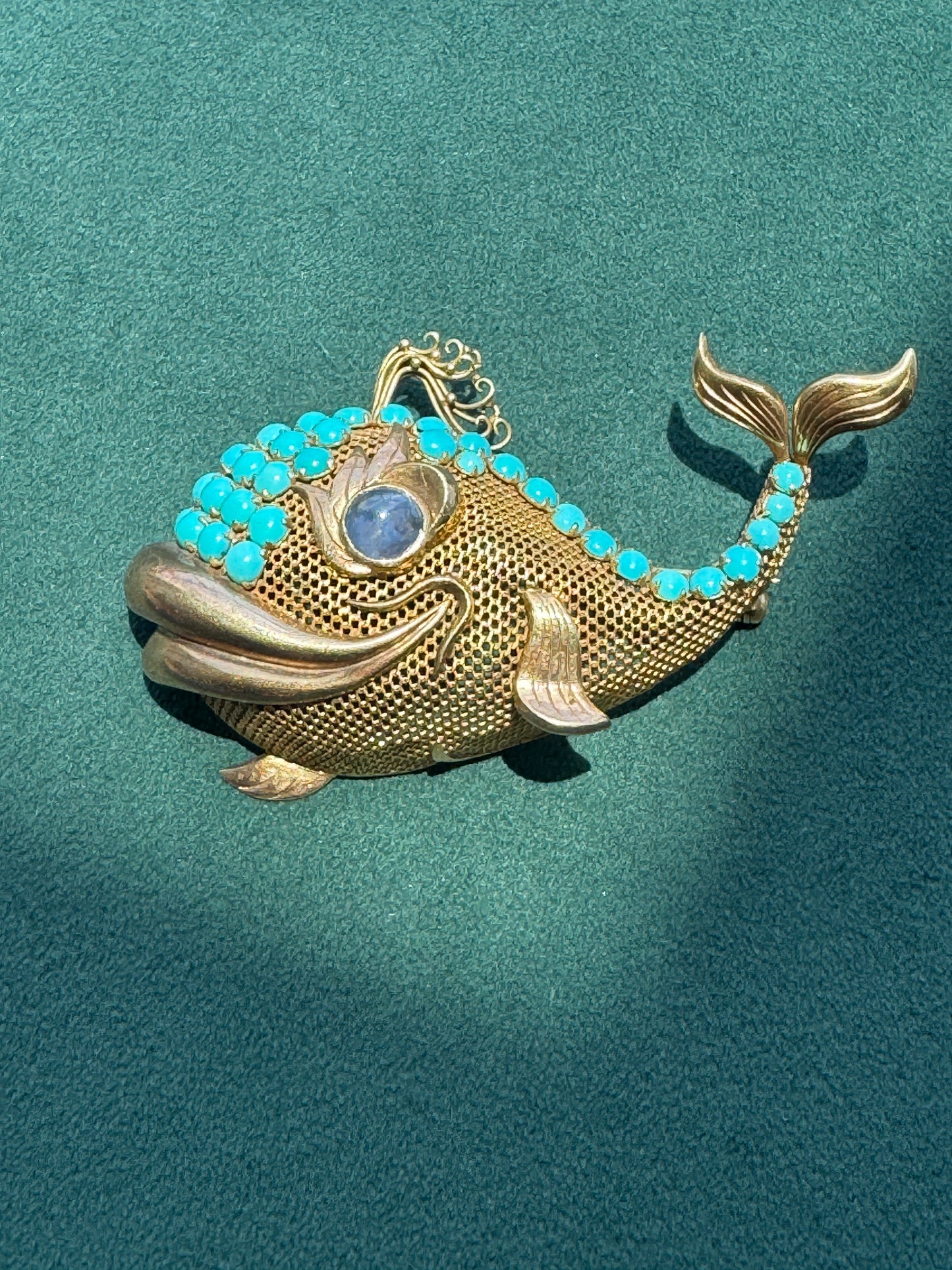 A gold fish-shaped brooch with turquoise blue rhinestone detailing and a blue cabochon stone for the eye, displayed on a textured teal background.