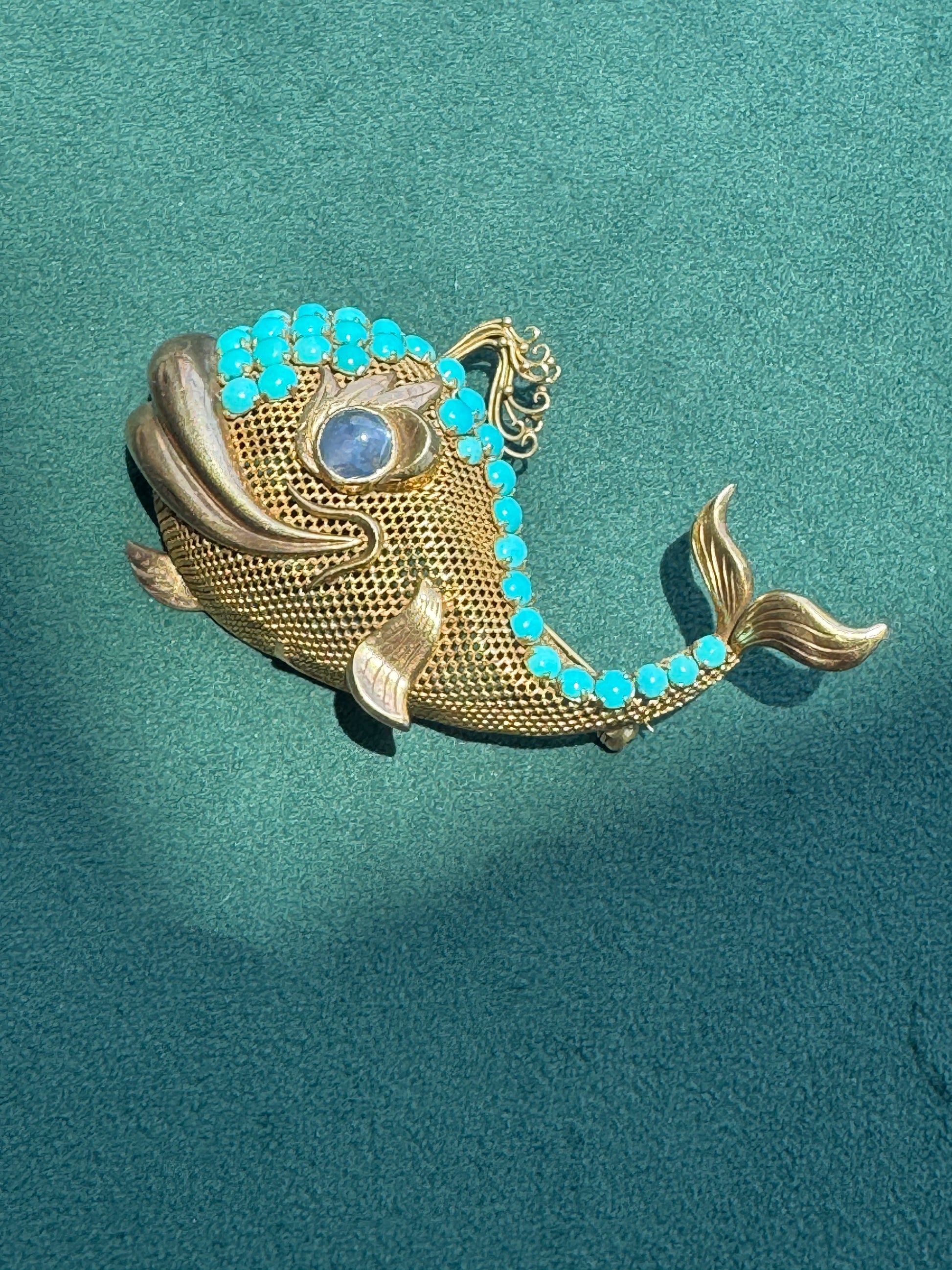 A gold fish-shaped brooch decorated with small turquoise blue stones and a blue oval stone for the eye, displayed on a textured teal background.