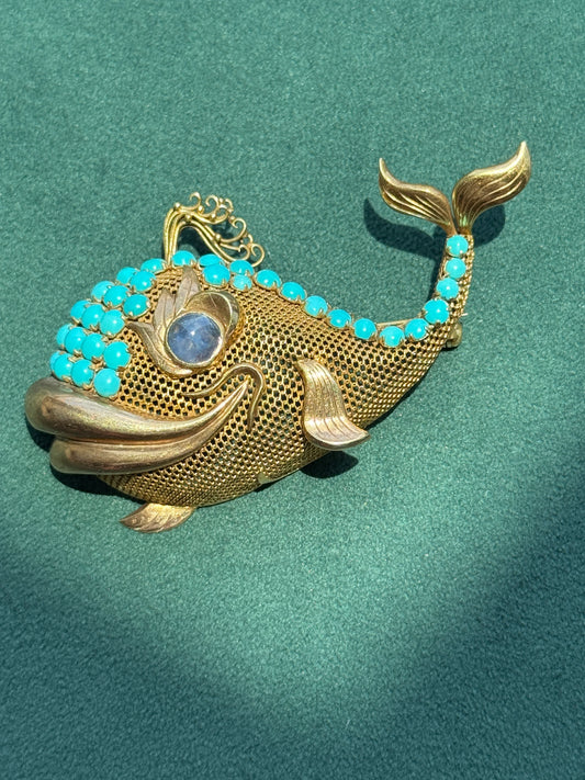 A gold fish-shaped brooch decorated with turquoise blue stones along the back and gills, featuring a round blue stone for the eye, displayed on a textured green background.