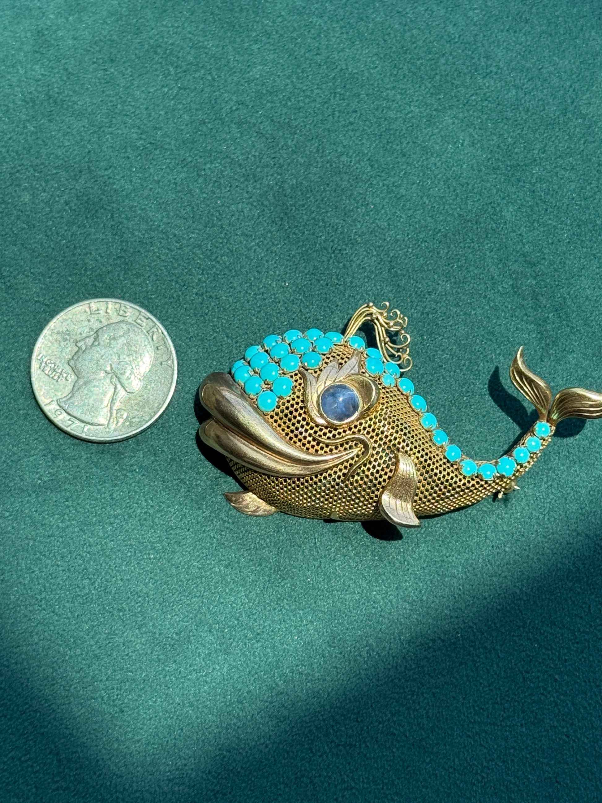 A gold fish-shaped brooch decorated with turquoise blue stones, shown next to a US quarter for scale on a dark green textured background.