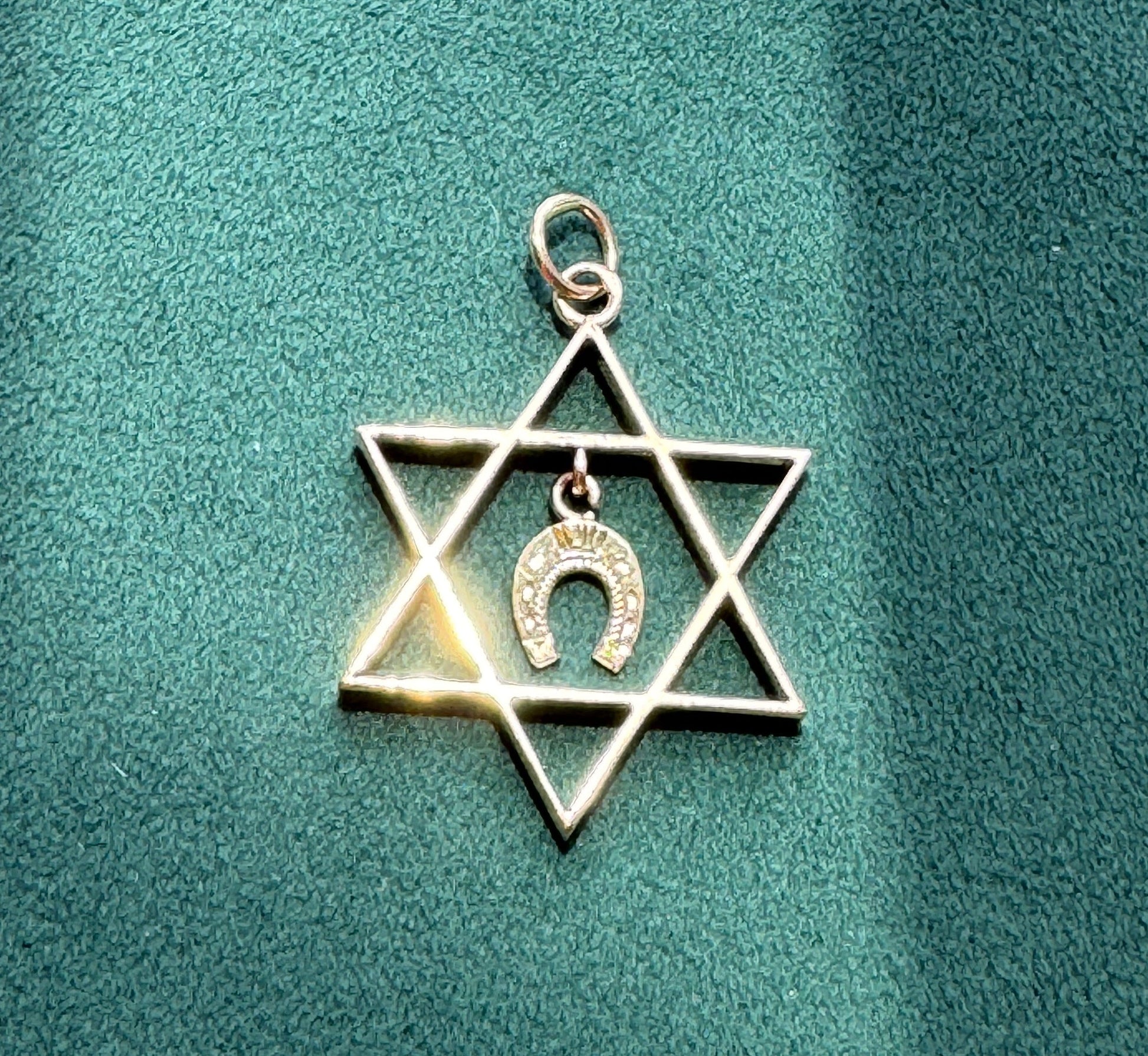 A silver-toned Star of David pendant with a small horseshoe charm in the center, displayed on a textured teal-green fabric background.