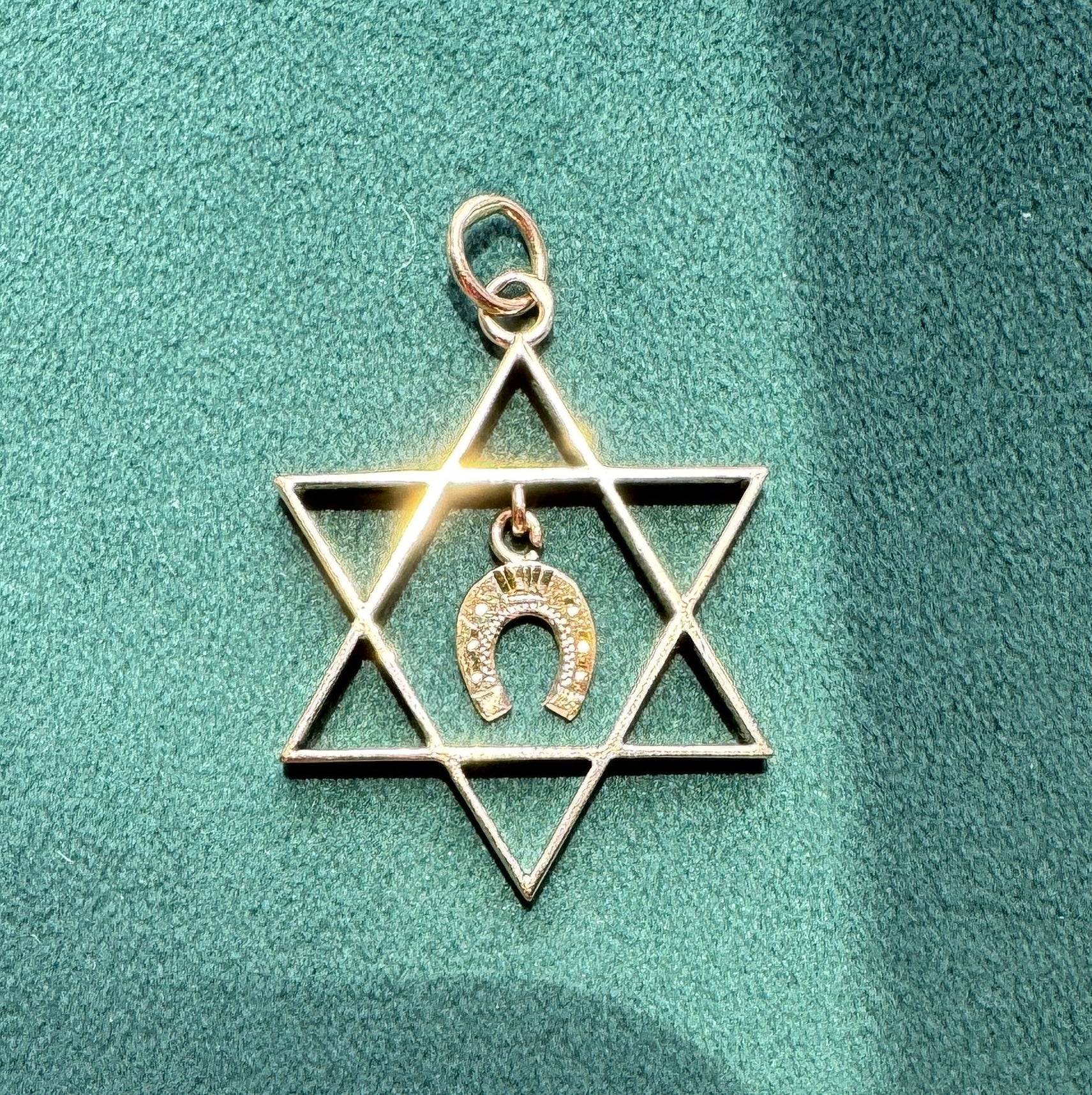 A gold-toned metal charm shaped like a Star of David with a small horseshoe pendant in the center, resting on a textured teal-green surface.