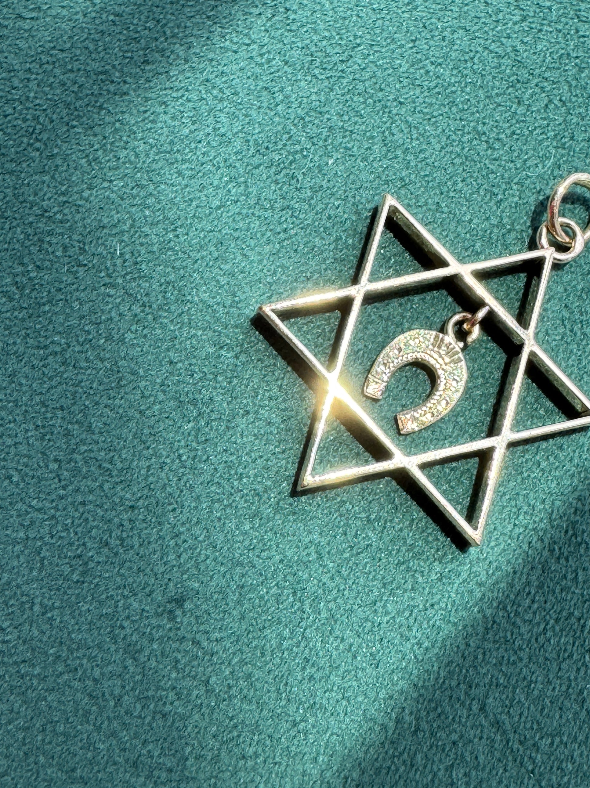 A silver-toned Star of David pendant with a small Shema letter charm, displayed on a textured teal fabric background.