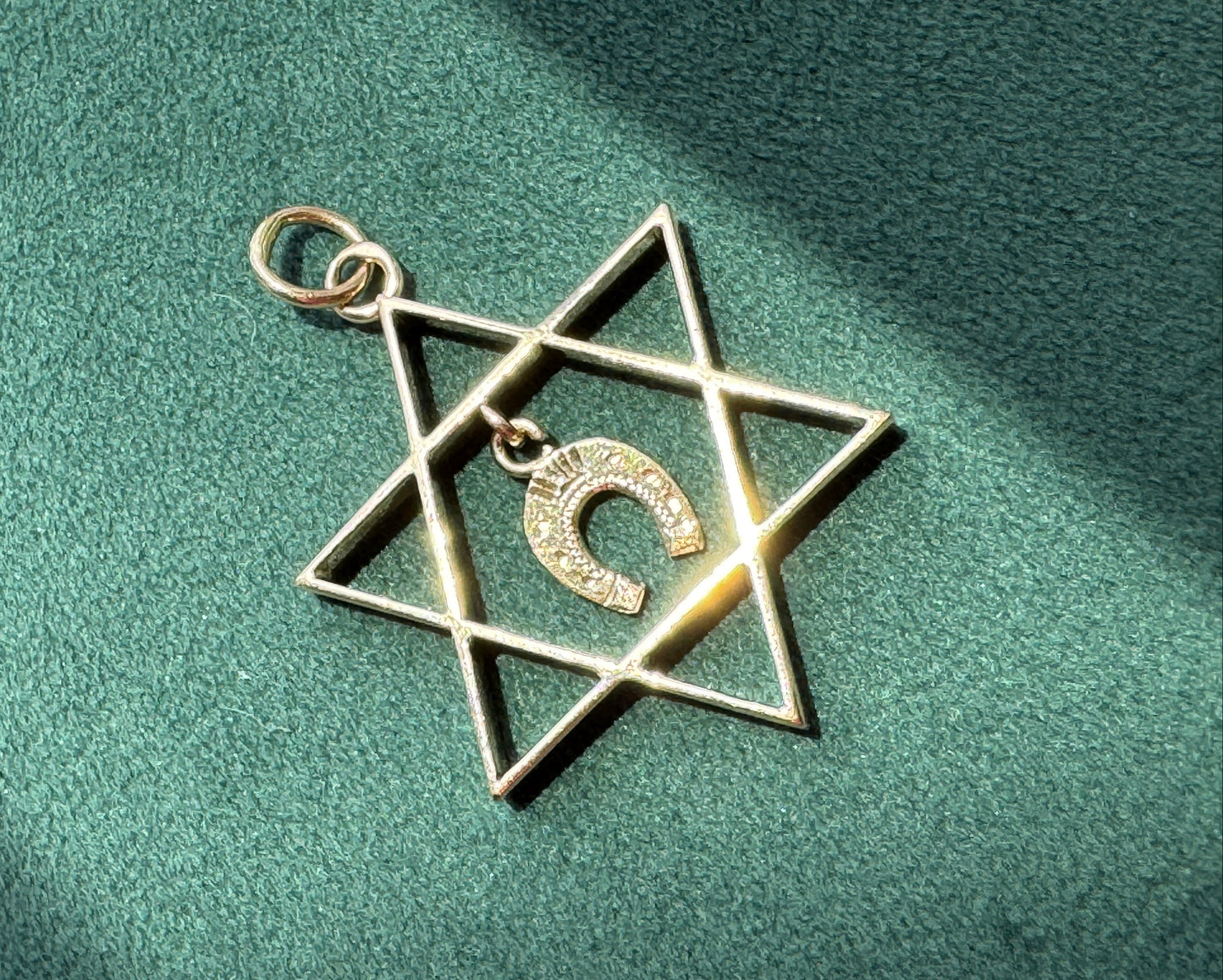 A gold-toned Star of David pendant with a small Hebrew letter charm in the center, resting on a textured green surface with soft shadows.