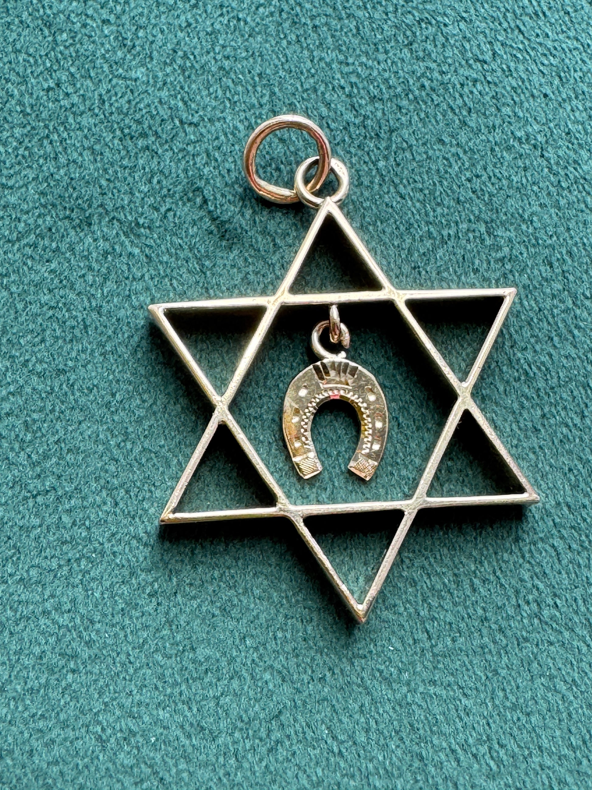 A silver-tone Star of David pendant featuring a small hanging horseshoe charm with a crown detail, displayed on a textured teal background.