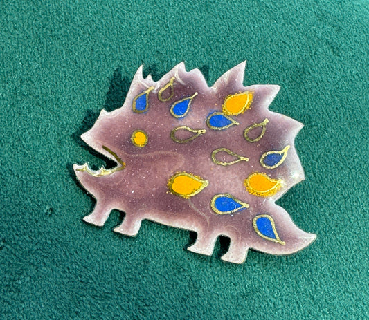 A dinosaur-shaped enamel pin in dusty purple with yellow and blue accents, displayed on a textured green surface.