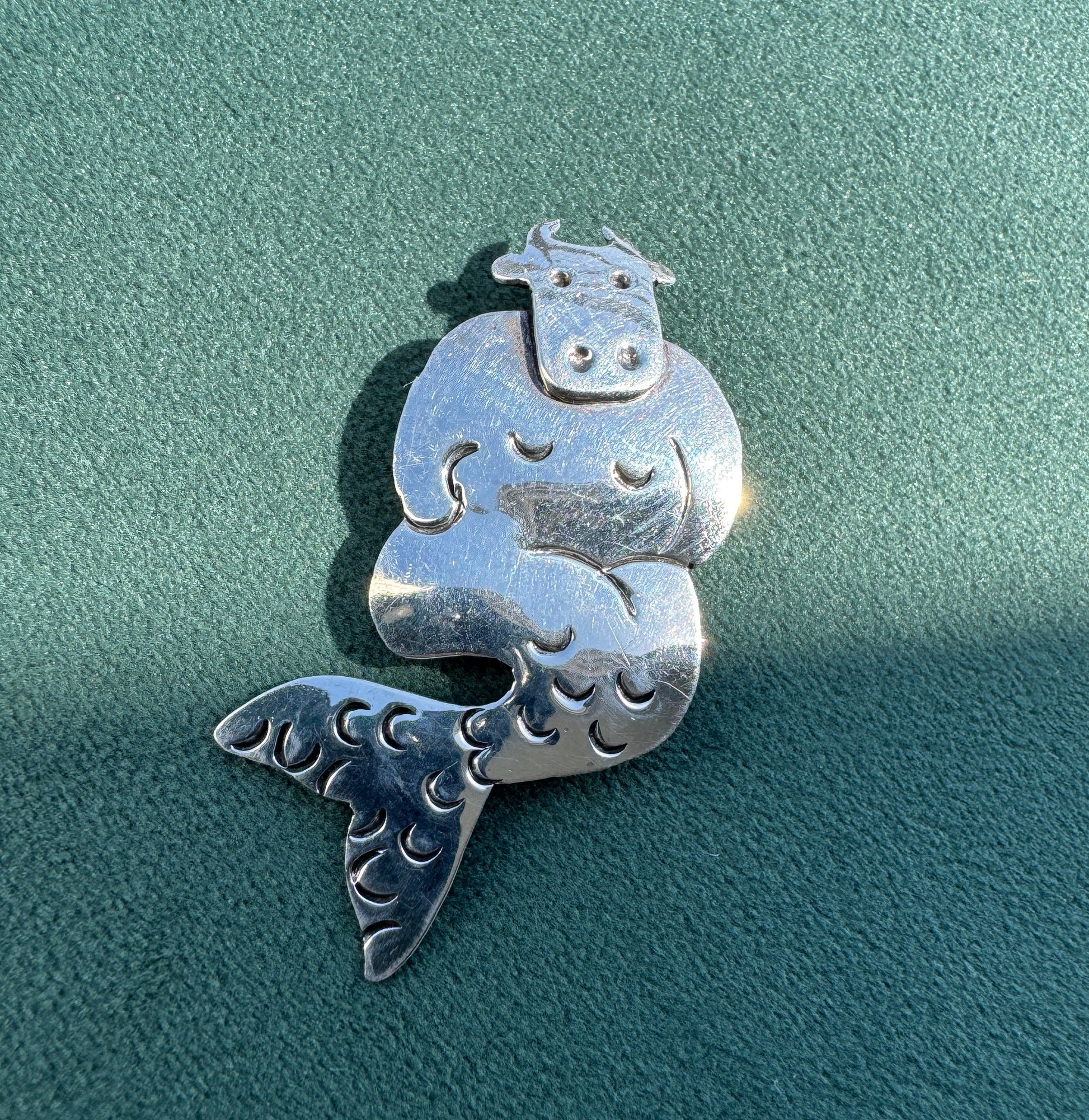 A sterling silver cow-and-alfalfa mermaid pendant with detailed hand-carved textures, displayed on a textured teal-green surface.
