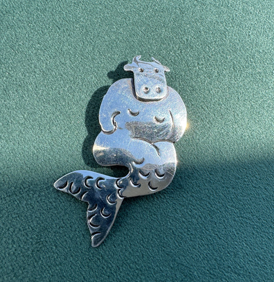 A sterling silver cow-and-alfalfa mermaid pendant with detailed hand-carved textures, displayed on a textured teal-green surface.