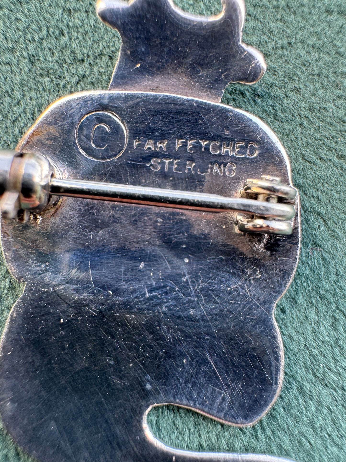 Close-up of the back of a silver fairy-shaped pin or brooch on a textured green background. The metal shows engraved text reading 'C FAN FEYCHED STERLING' and a butterfly clutch fastener.