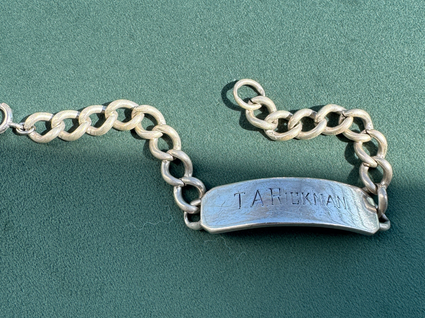 A silver nameplate bracelet with a textured, engraved plate connected to a curb chain, displayed on a dark green textured background.