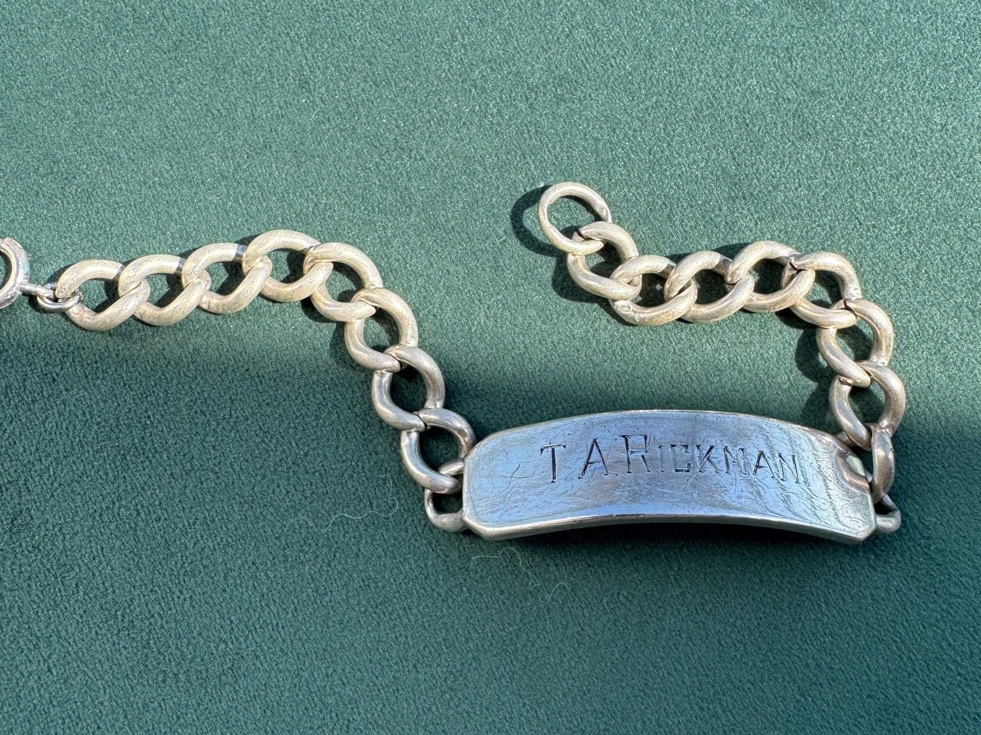 A silver nameplate bracelet with a textured, engraved plate connected to a curb chain, displayed on a dark green textured background.