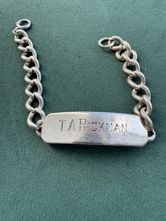 A silver-tone metal chain bracelet with a rectangular nameplate engraved with the name 'T.A.