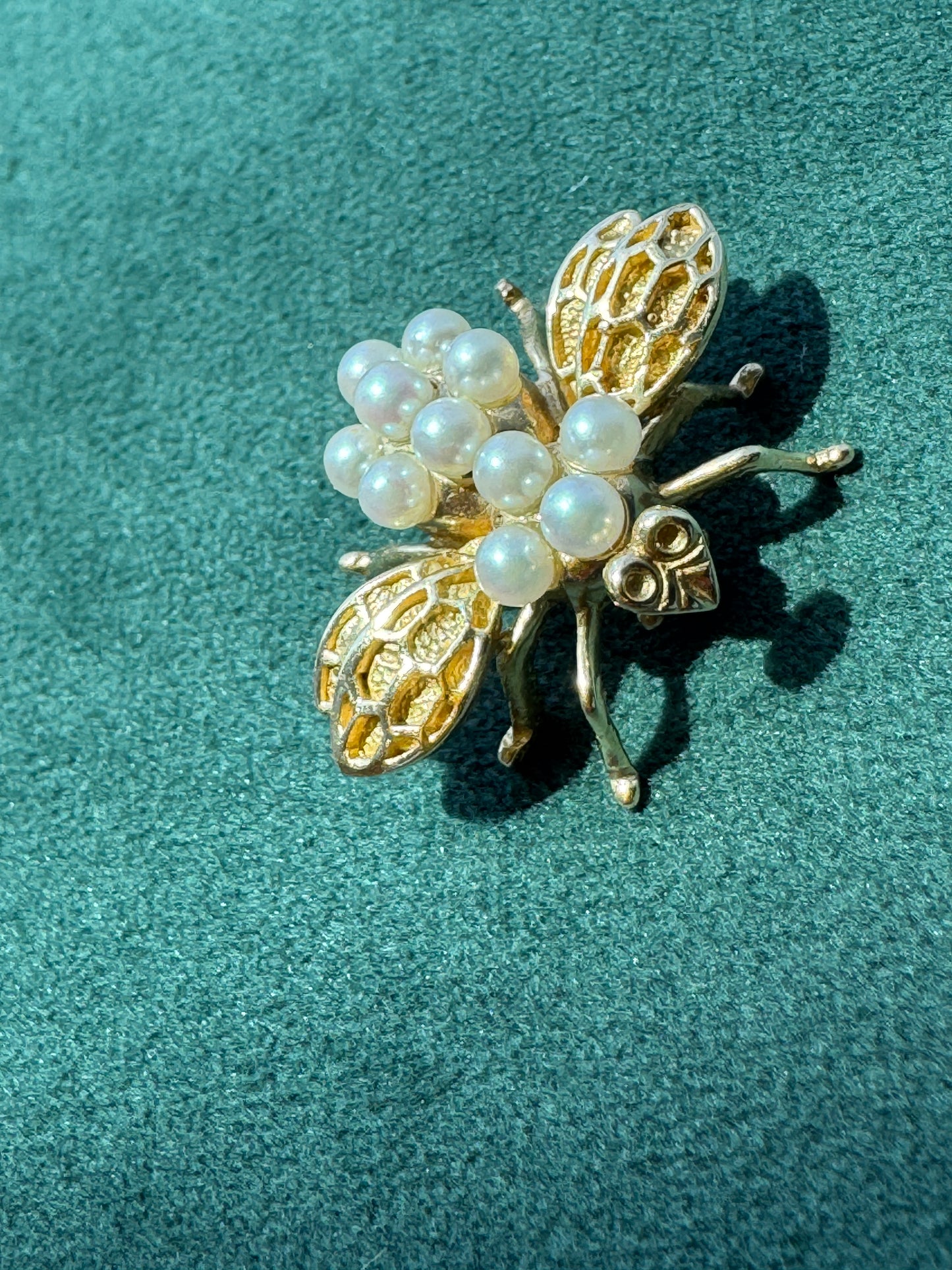 A gold insect brooch designed like a bee or beetle, adorned with white faux pearls, resting on a textured teal-green fabric background.