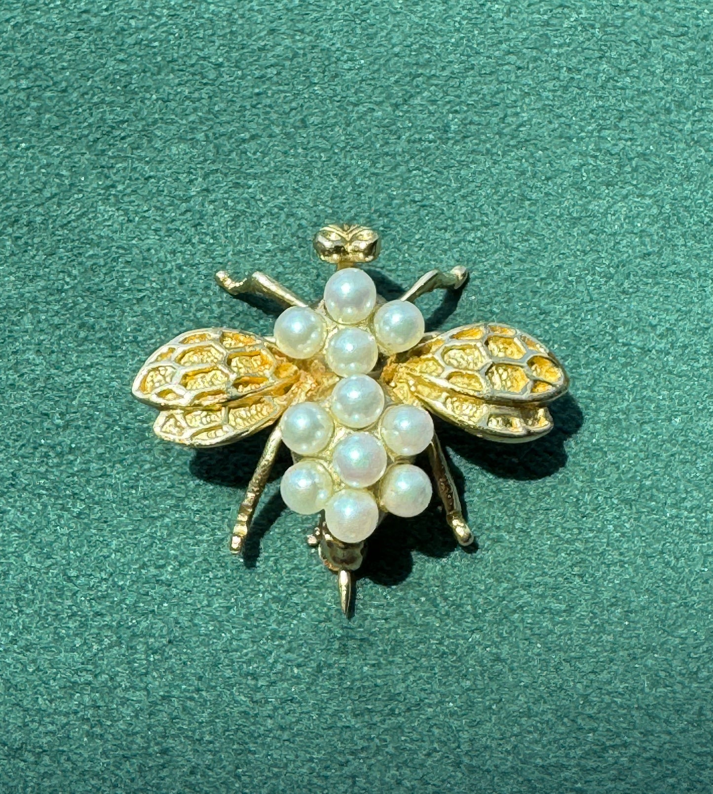 A gold bee-shaped brooch embellished with white faux pearls, displayed on a textured dark green background.