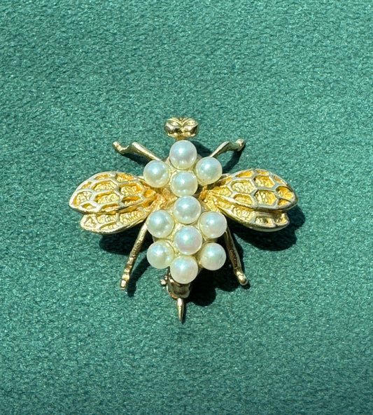 A gold bee-shaped brooch embellished with white faux pearls, displayed on a textured dark green background.