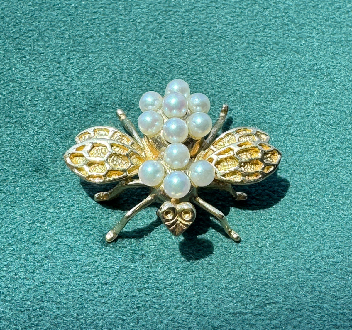A gold-tone bee brooch embellished with white faux pearls, displayed on a textured teal-green background.