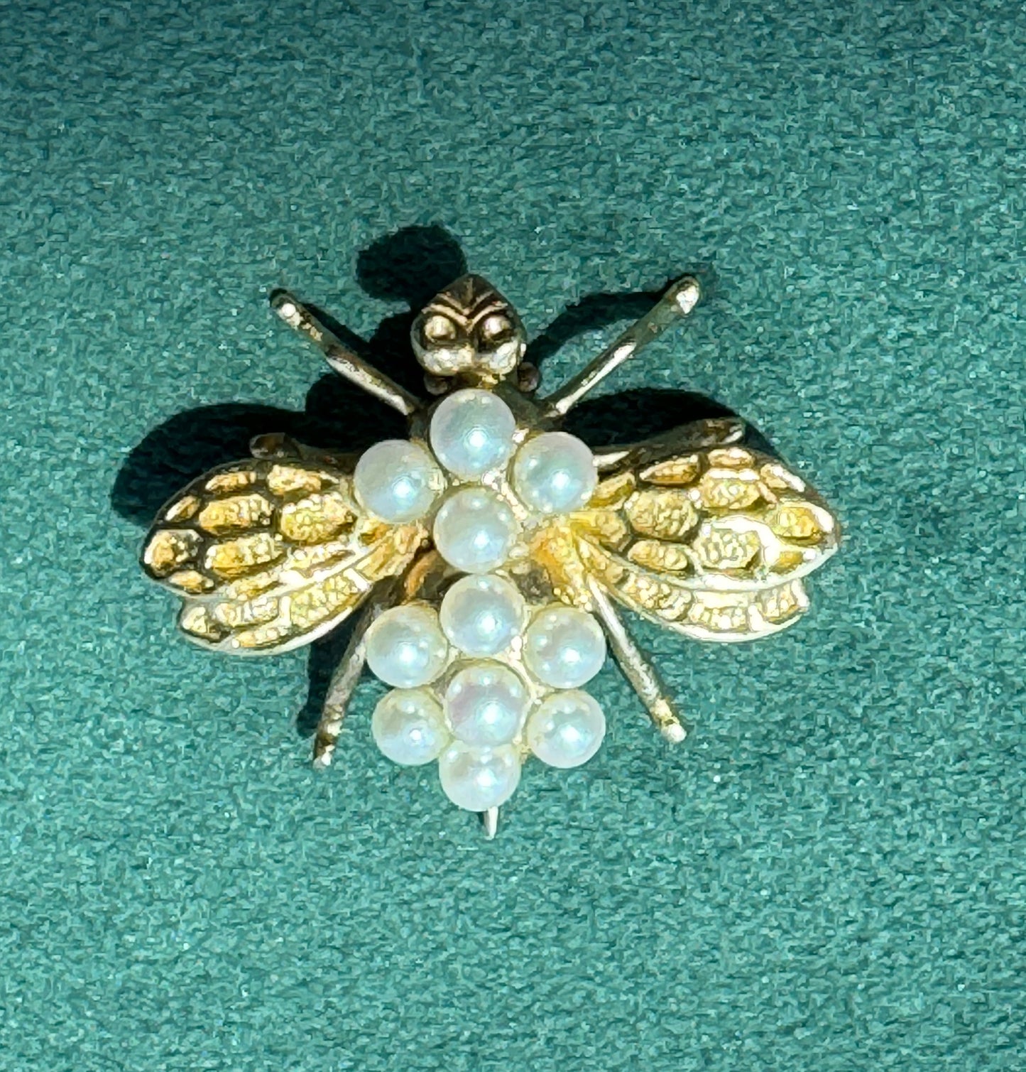 A vintage-style bee brooch featuring textured gold-tone wings and legs with a cluster of white faux pearls forming the body, displayed on a textured teal-green background.