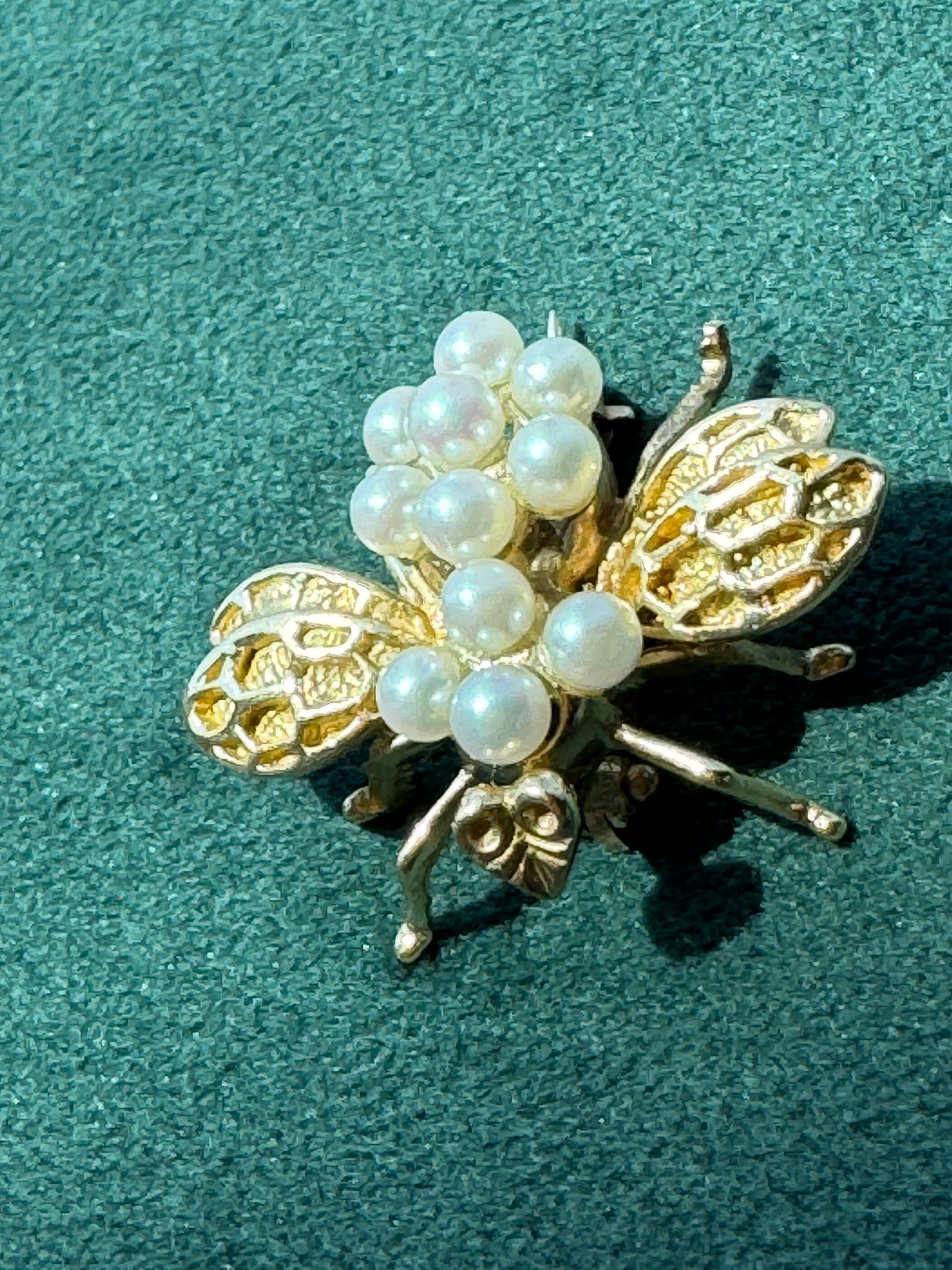 A gold-tone bee brooch embellished with white faux pearls, displayed on a textured dark green background.