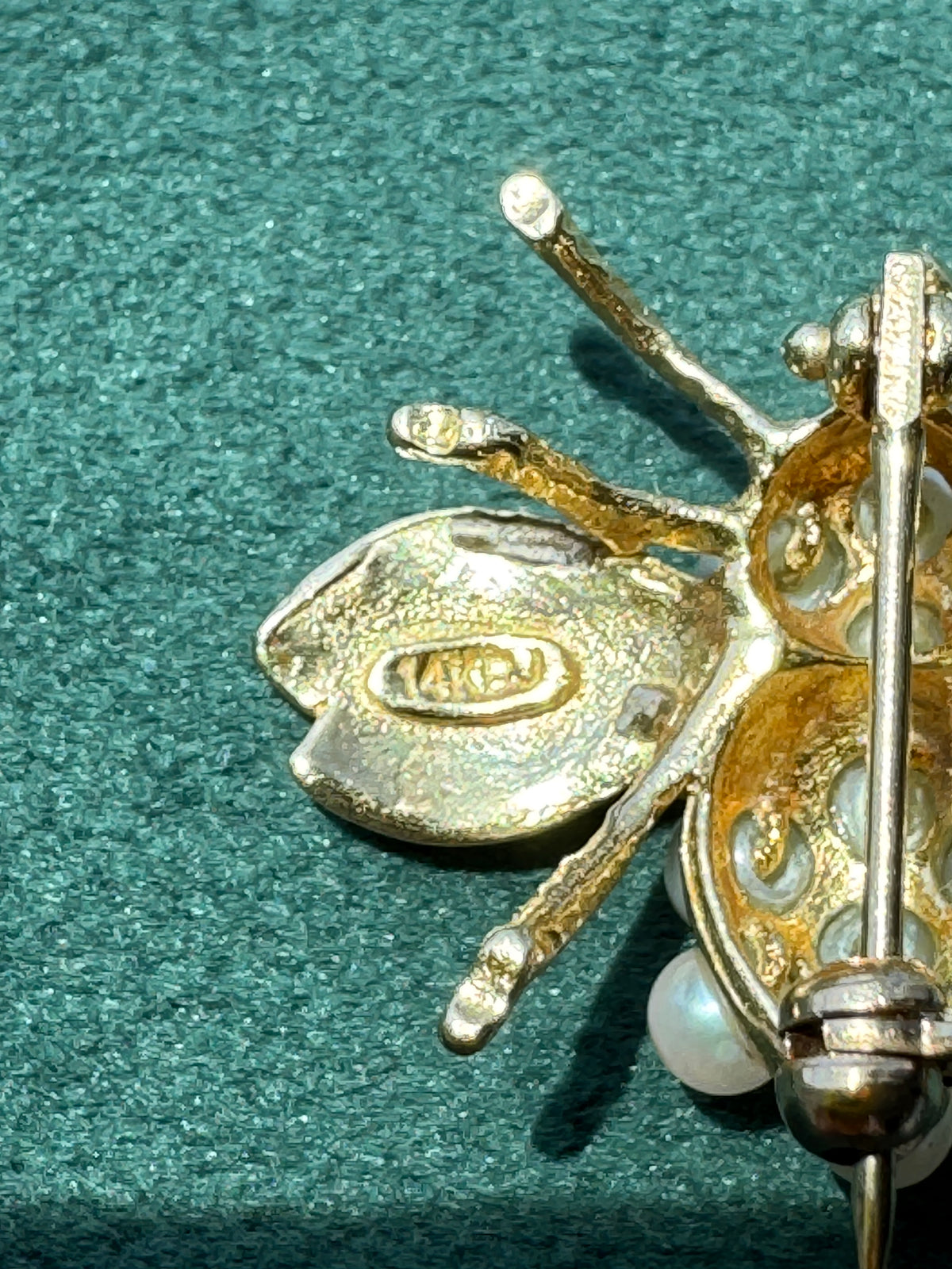 Close-up of a gold-toned insect brooch, likely a beetle or spider, with a pearl accent, resting on a textured dark green surface. The back of the wing features a stamped brand logo reading 'MARC'.