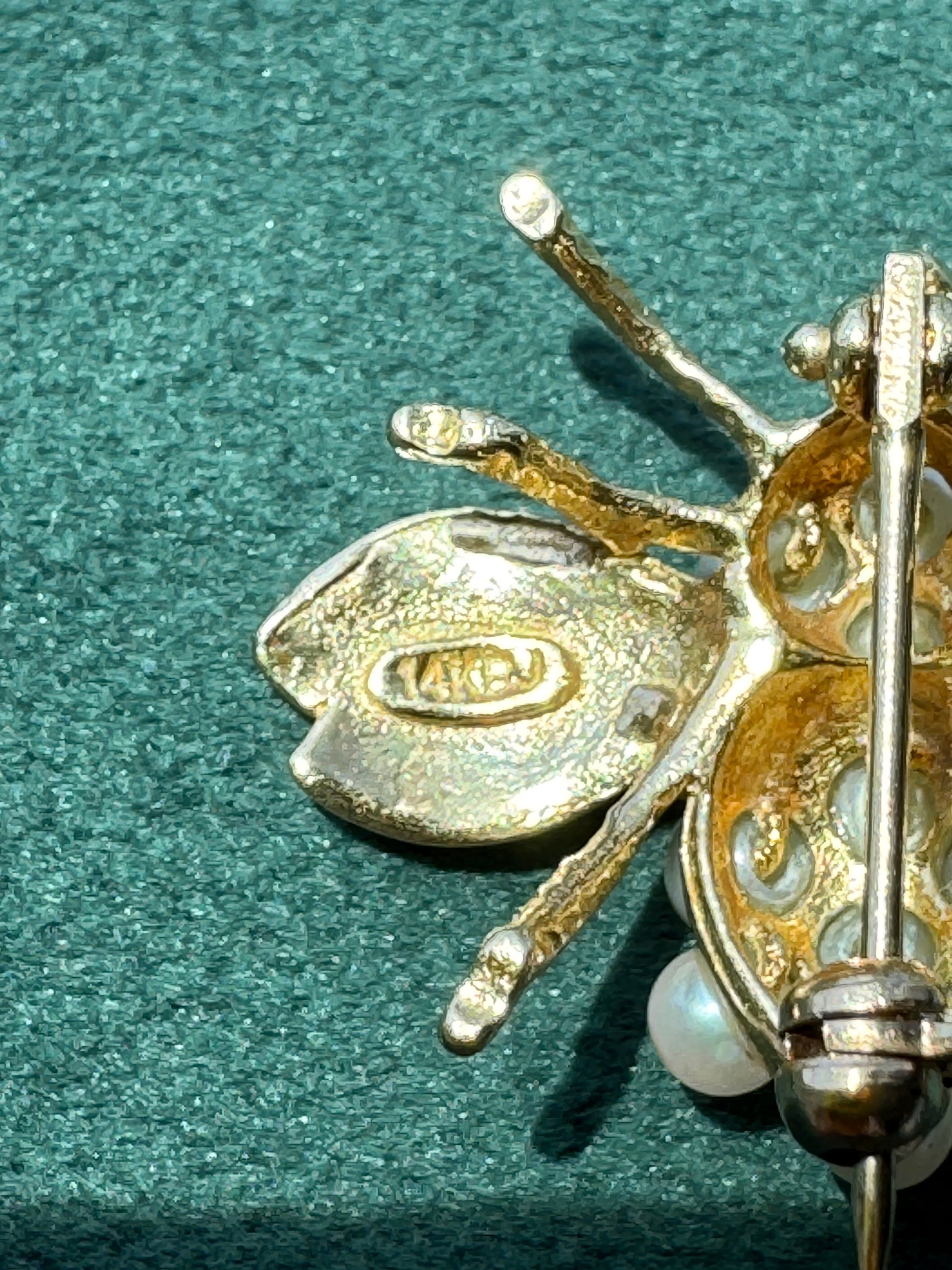 Close-up of a gold-toned insect brooch, likely a beetle or spider, with a pearl accent, resting on a textured dark green surface. The back of the wing features a stamped brand logo reading 'MARC'.