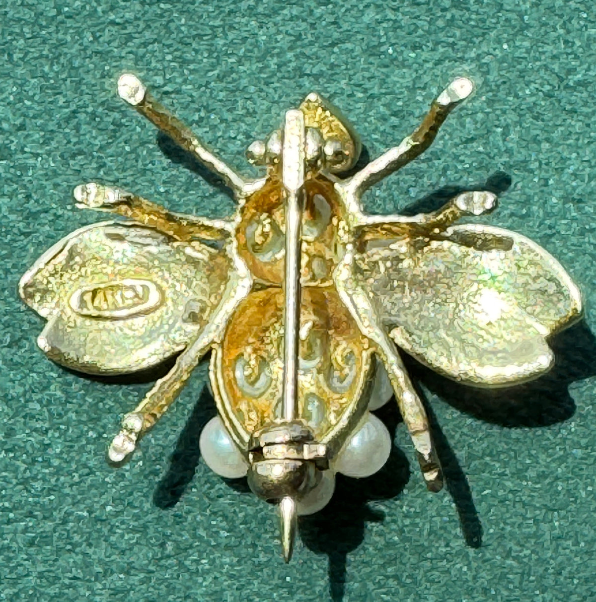 A gold and silver bee-shaped brooch with pearl accents, displayed on a textured green surface.
