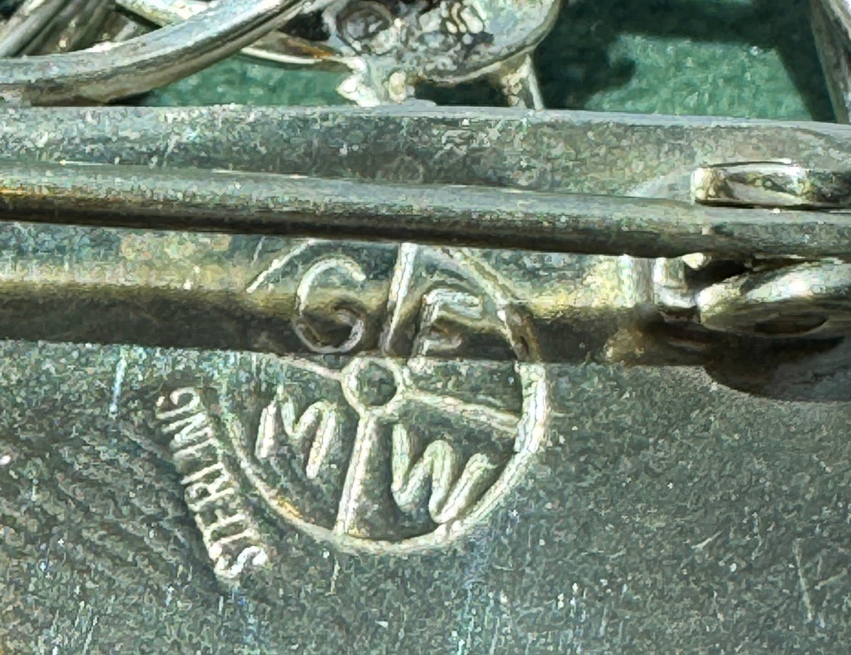 Close-up of a silver-toned metal accessory with a pin fastener, engraved initials 'GM MW', and a small bird motif, shown against a textured green background.