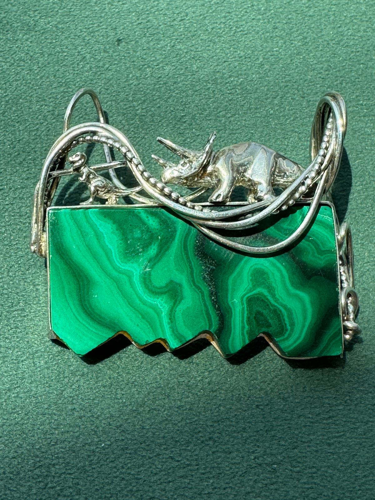 A silver-toned jewelry piece featuring a rectangular green malachite stone base with wavy cut edges, topped with a small silver triceratops figure and looping silver wire details, shown on a textured dark green background.