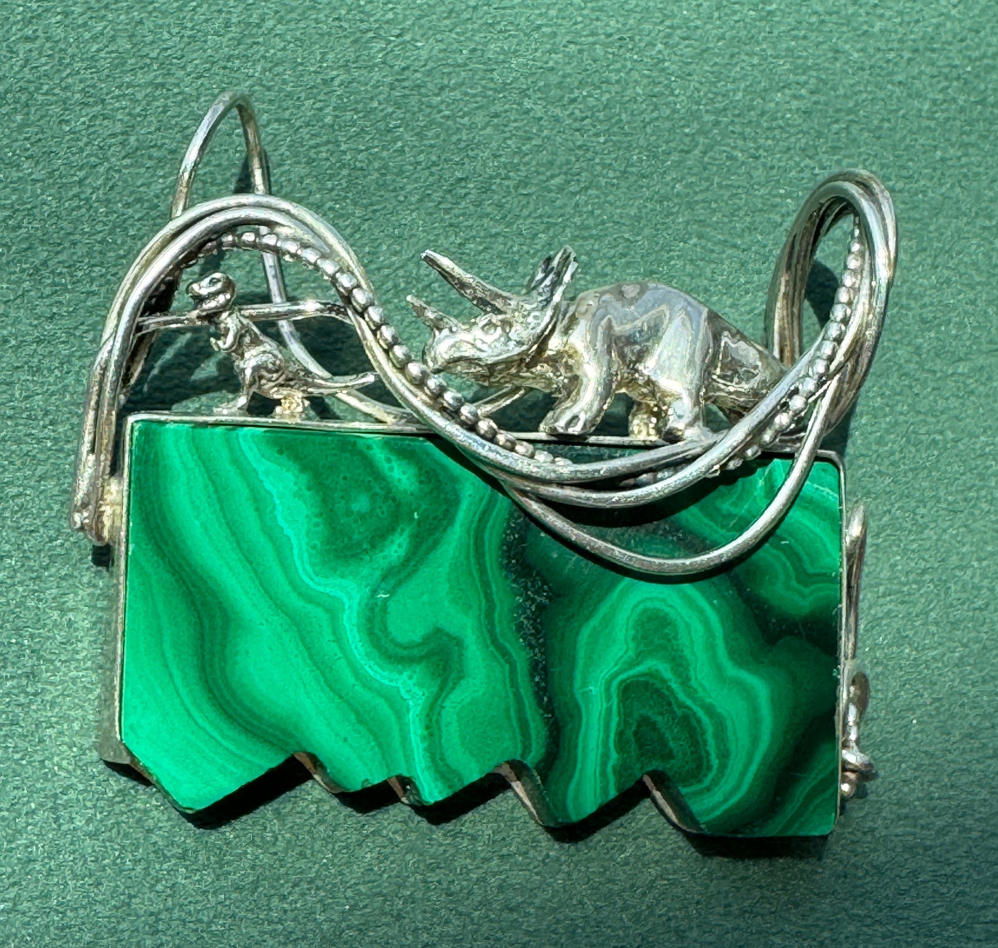 A silver-toned triceratops dinosaur pendant with a rectangular green malachite stone base, displayed on a textured green background.