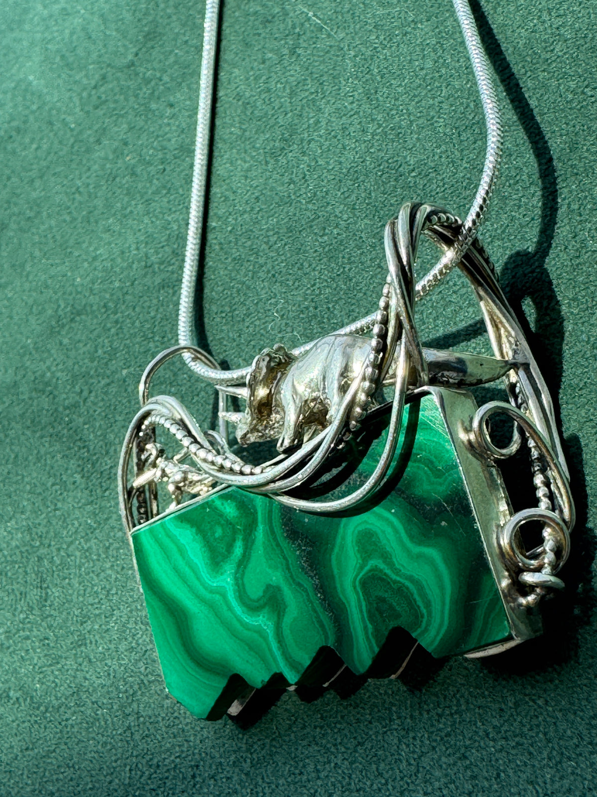A silver-toned necklace with a geometric pendant featuring a prominent green malachite stone, displayed on a textured green background.