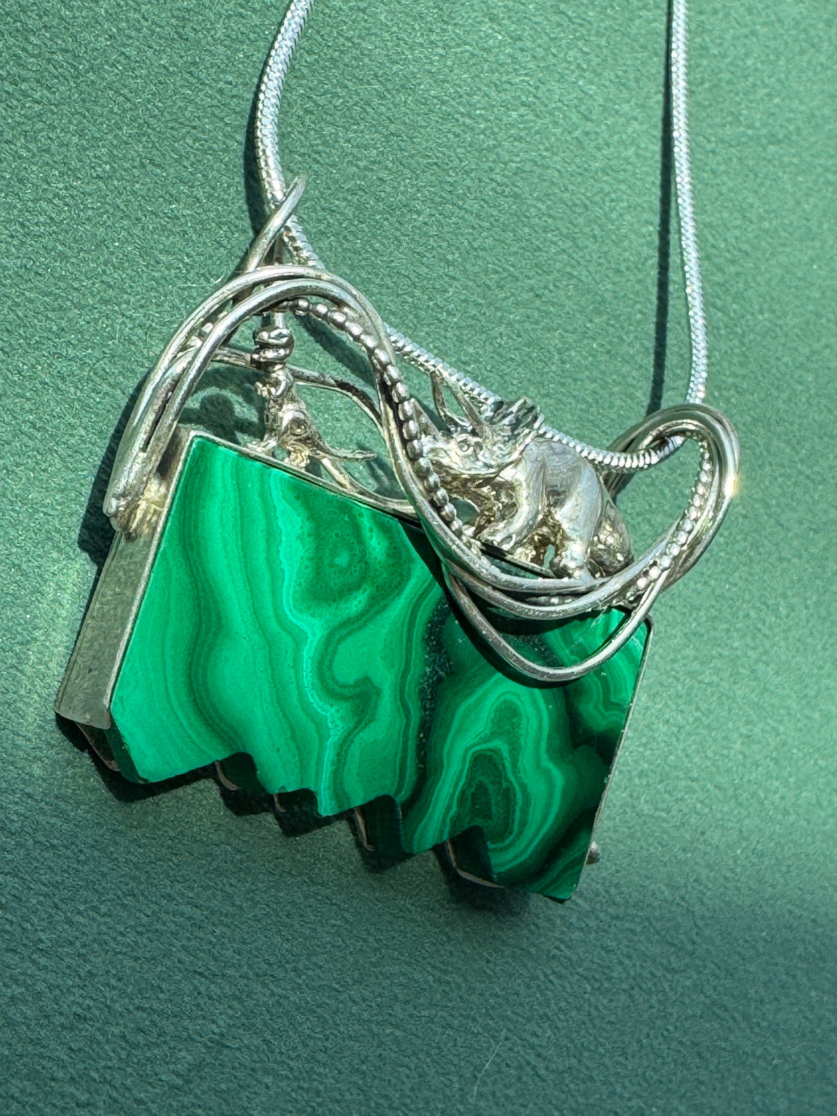 A silver-toned necklace featuring a unique pendant made of stacked, irregular green malachite stone slices, resting on a textured green background.