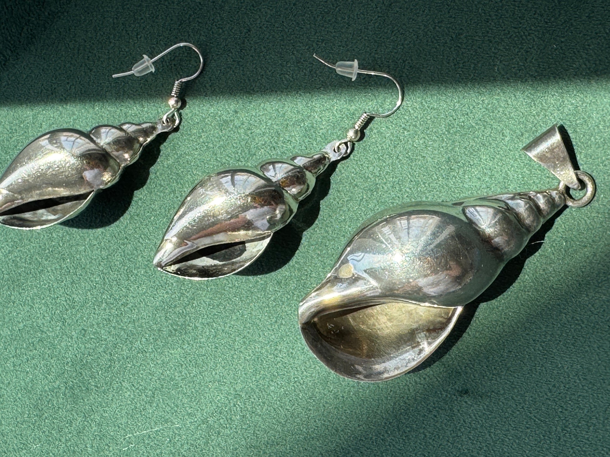 Three silver conch shell dangle earrings with fishhook backs, arranged diagonally on a textured green surface with hard sunlight shadows.