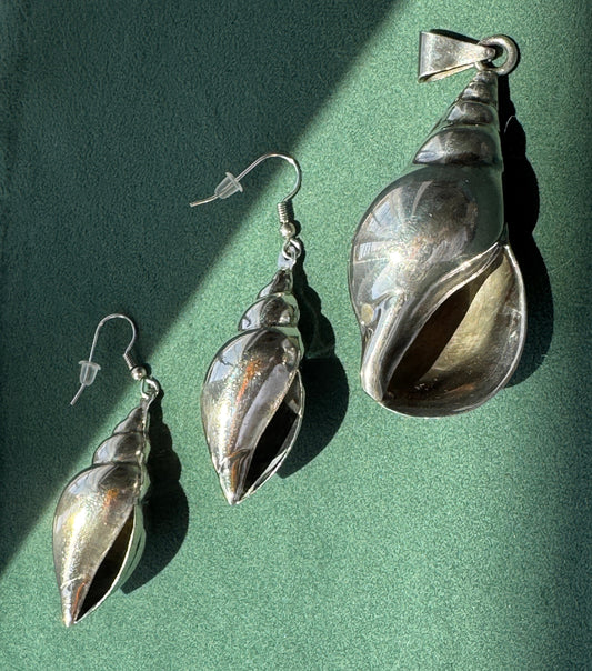 Three silver shell-shaped jewelry pieces arranged on a textured green surface: one large pendant, one matching drop earring, and one smaller drop earring. Strong directional sunlight casts sharp shadows across the scene.