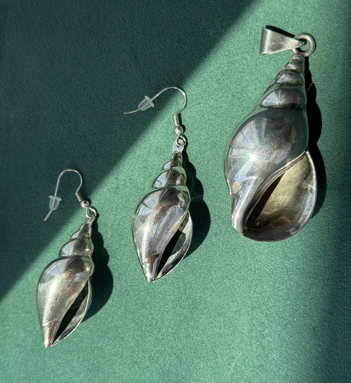 Three silver conch shell jewelry pieces, including two drop earrings and a matching pendant, arranged on a textured green surface with diagonal shadows.