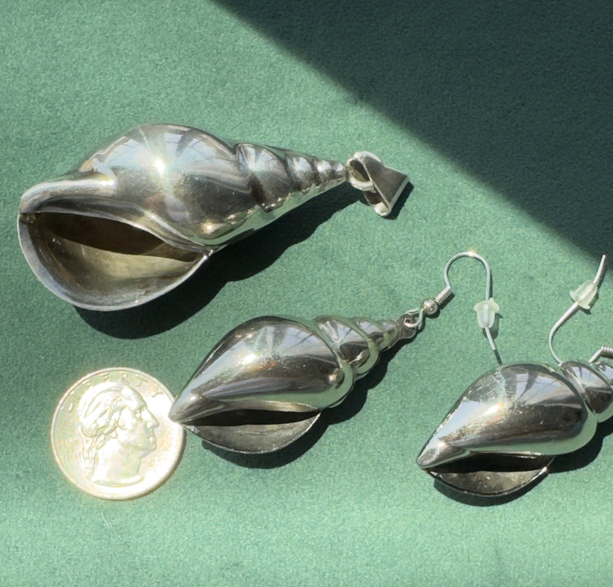 Three polished silver shell-shaped earrings displayed on a green surface, with a U.S.
