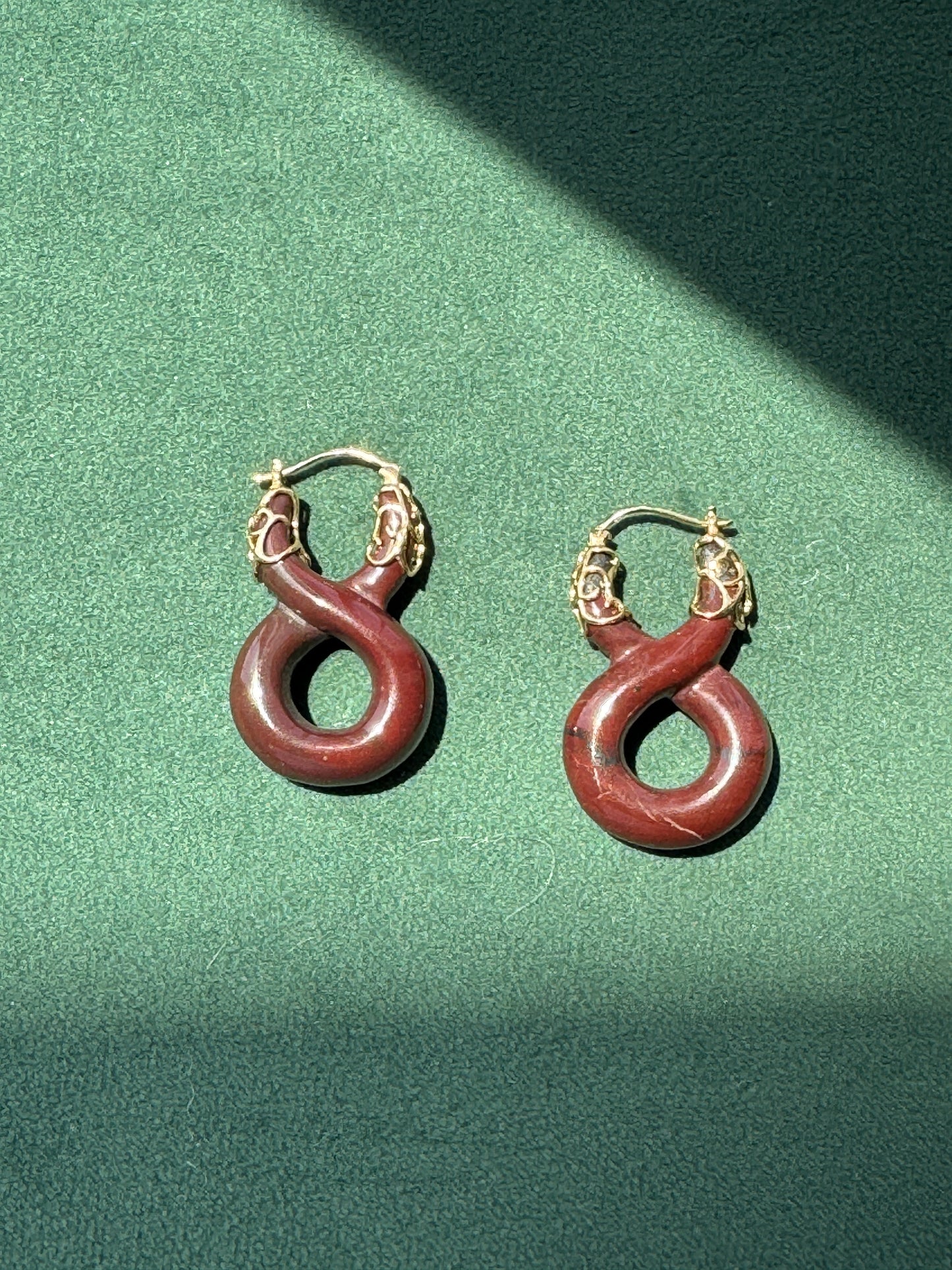 A pair of hoop earrings with reddish-brown coiled snake-shaped charms and gold-toned tops, displayed on a textured green surface with dramatic diagonal shadows.