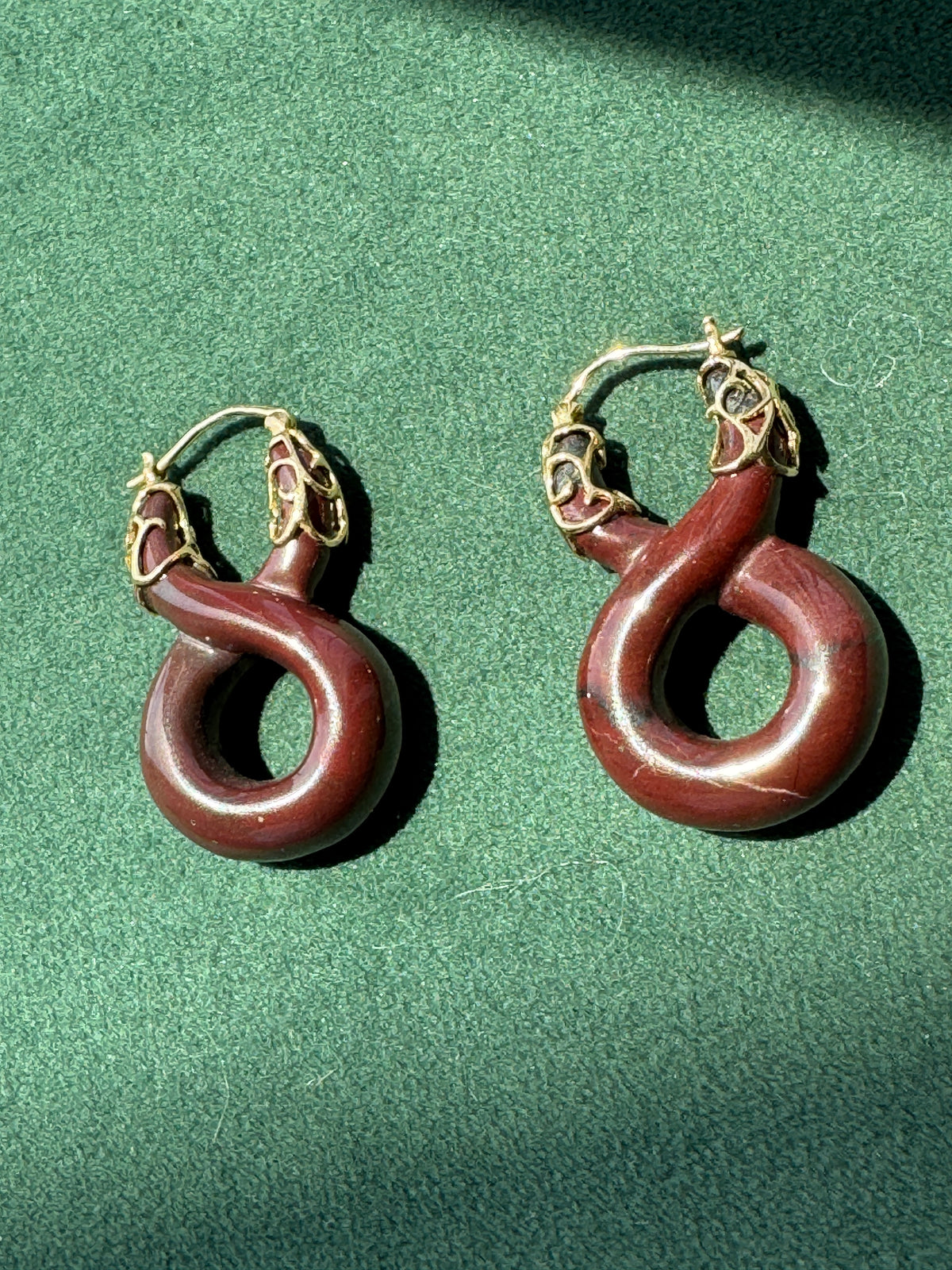A pair of hoop earrings with dark reddish-brown curved stone pendants and gold-toned metal hoops, displayed on a textured green surface.