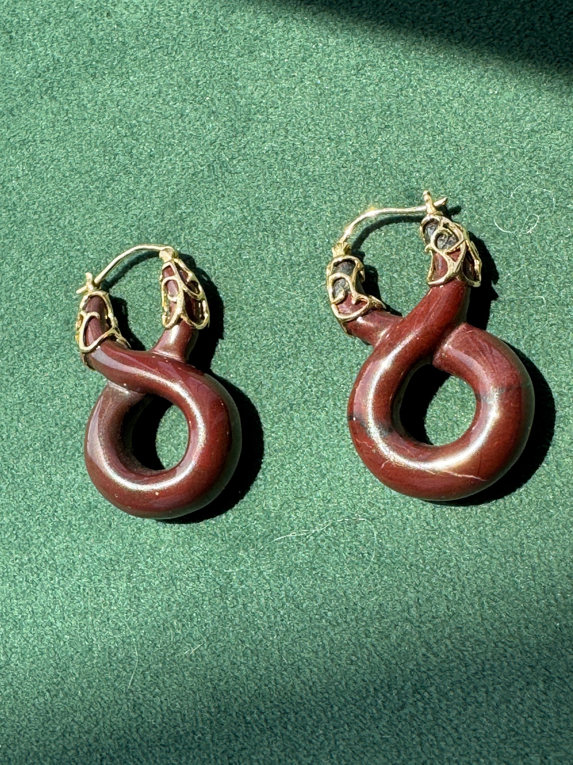 A pair of hoop earrings with dark reddish-brown curved stone pendants and gold-toned metal hoops, displayed on a textured green surface.
