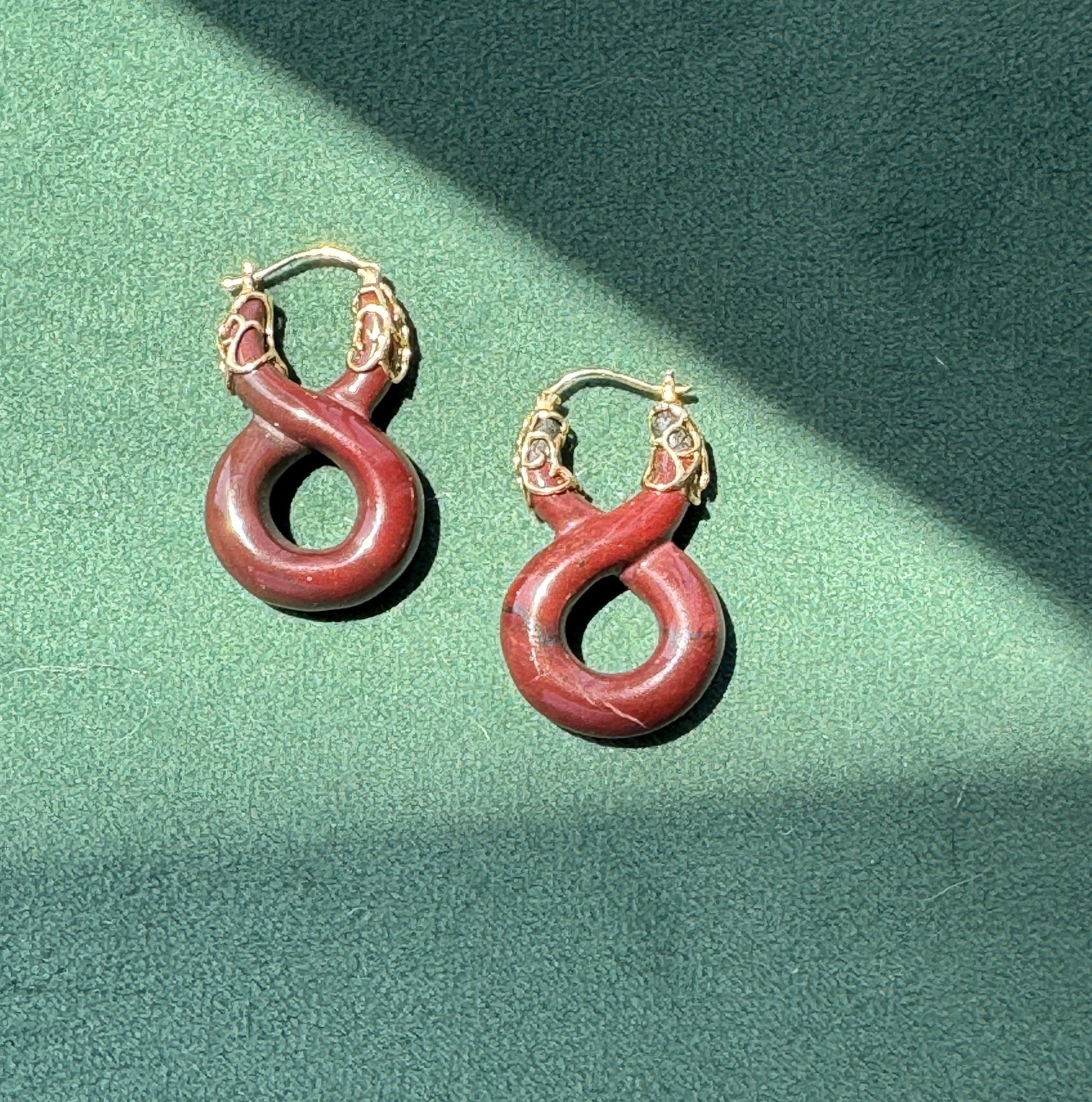 A pair of hoop earrings with reddish-brown circular drop charms and gold-toned hoops, displayed on a textured green surface with diagonal shadows.