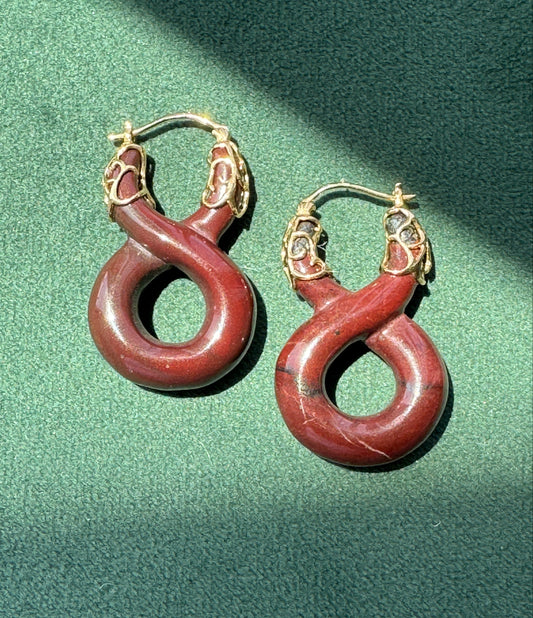 A pair of gold hoop earrings with large, coiled red stone pendants, displayed on a textured green surface with dramatic diagonal shadows.