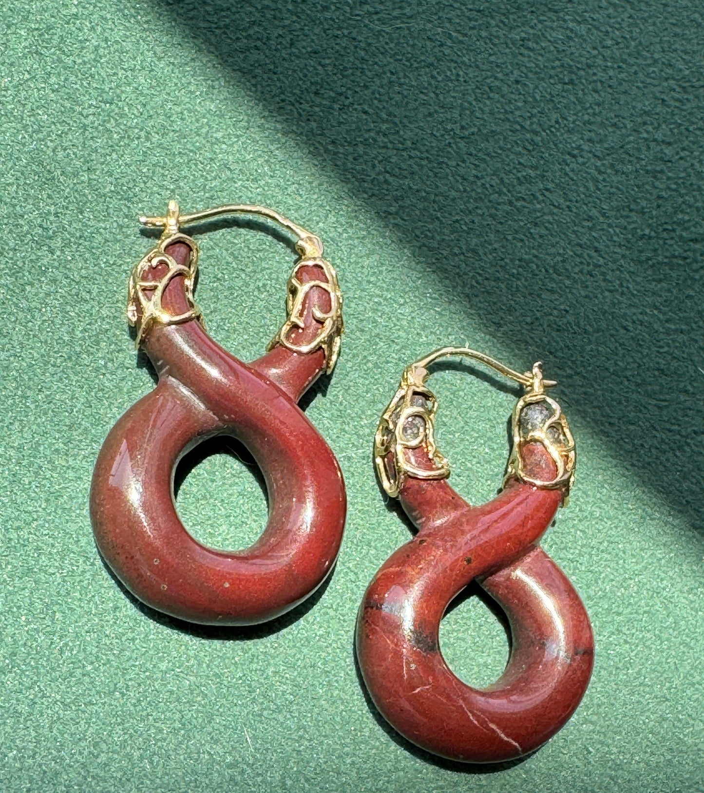 A pair of hoop earrings with carved red stone loops and gold-toned head details, displayed on a textured green surface.