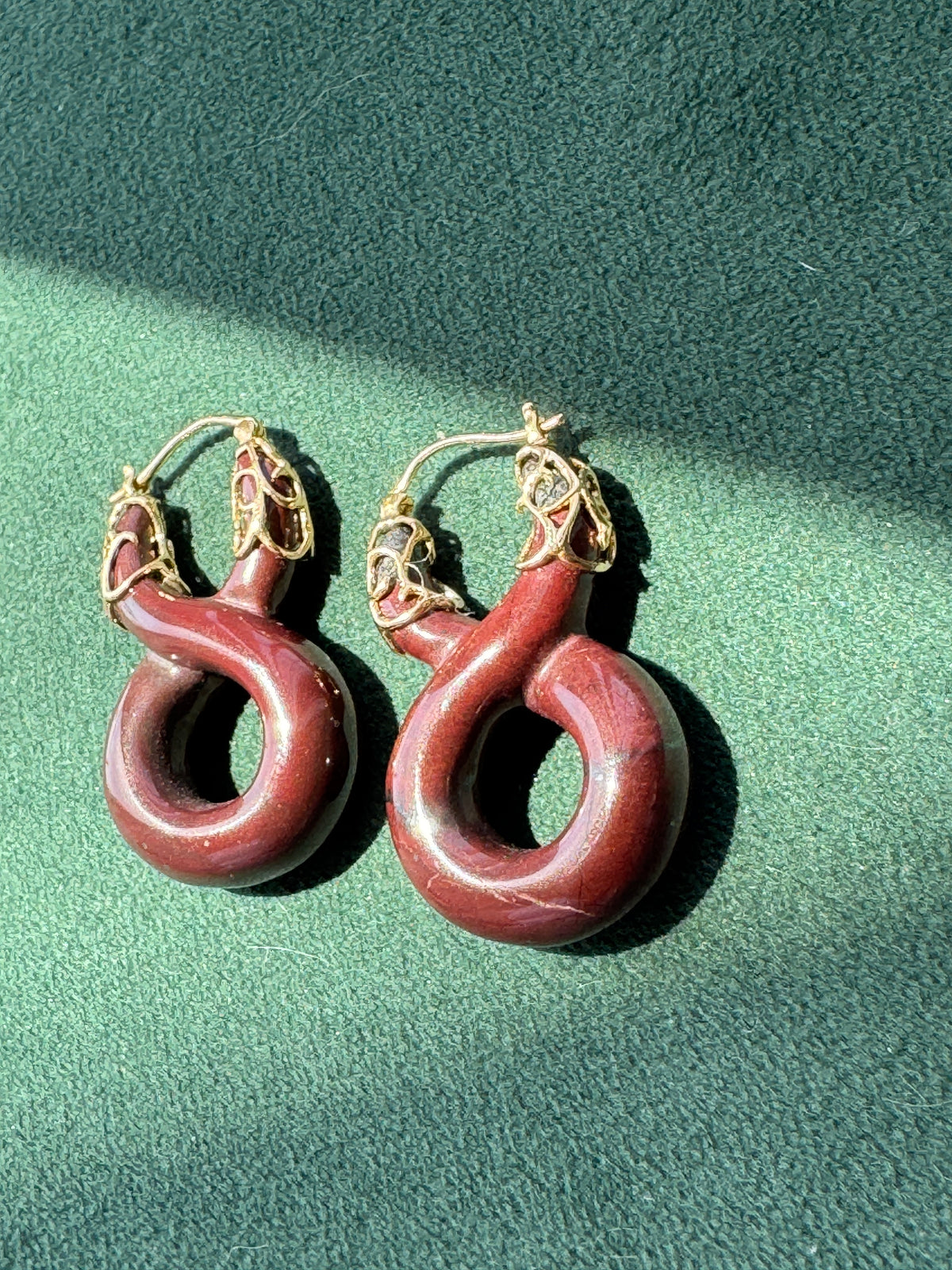A pair of large, coiled hoop earrings made from reddish-brown wood with gold-toned metal wire wrapping at the top, displayed on a textured green surface under direct sunlight.