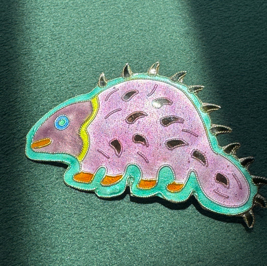 A colorful embroidered patch shaped like a stylized dinosaur or creature, shown on a dark green textured background.