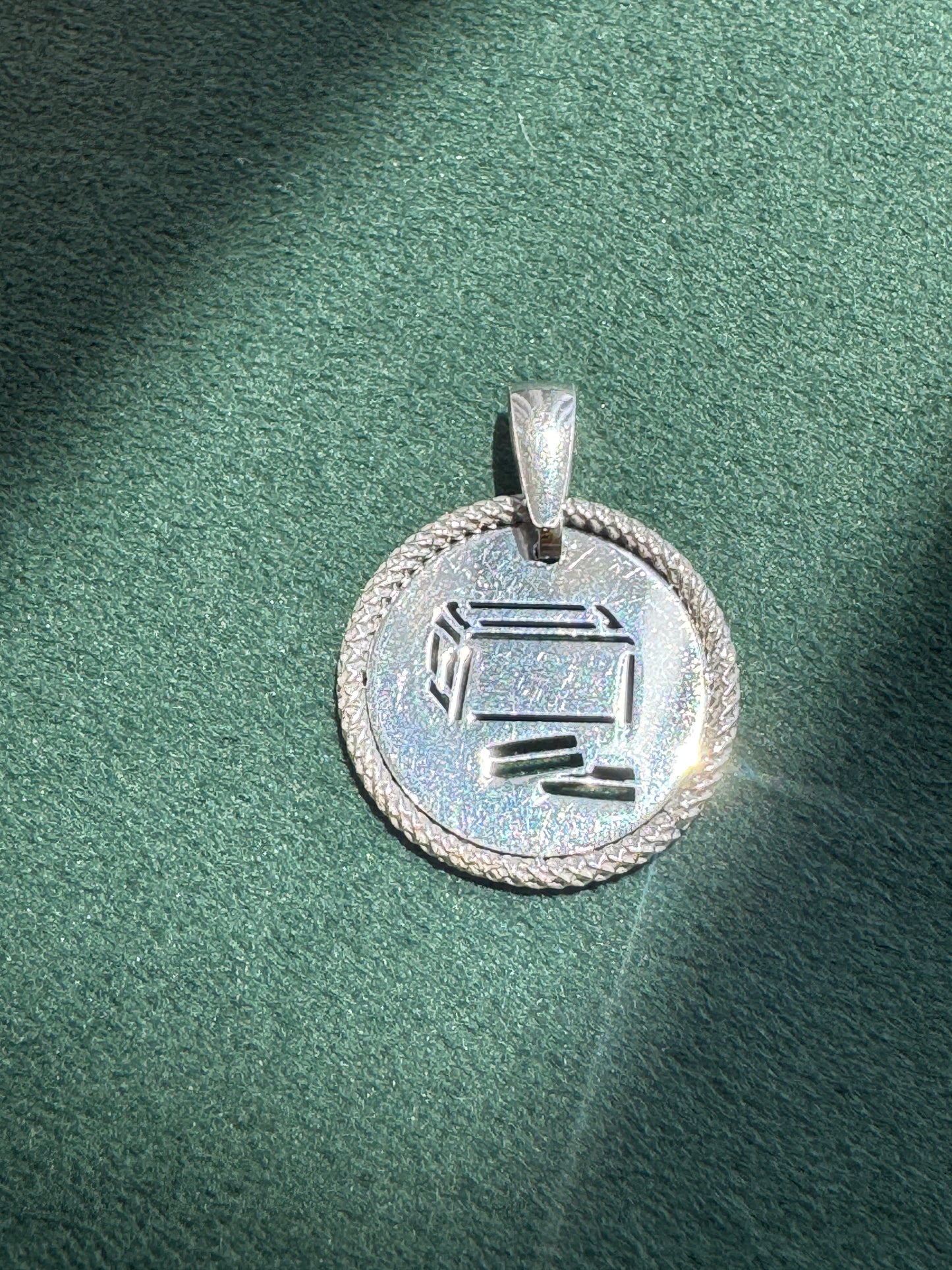 A silver pendant with a circular design, featuring a Hebrew letter 'Shin' engraved in the center and a braided border, resting on a textured green surface.