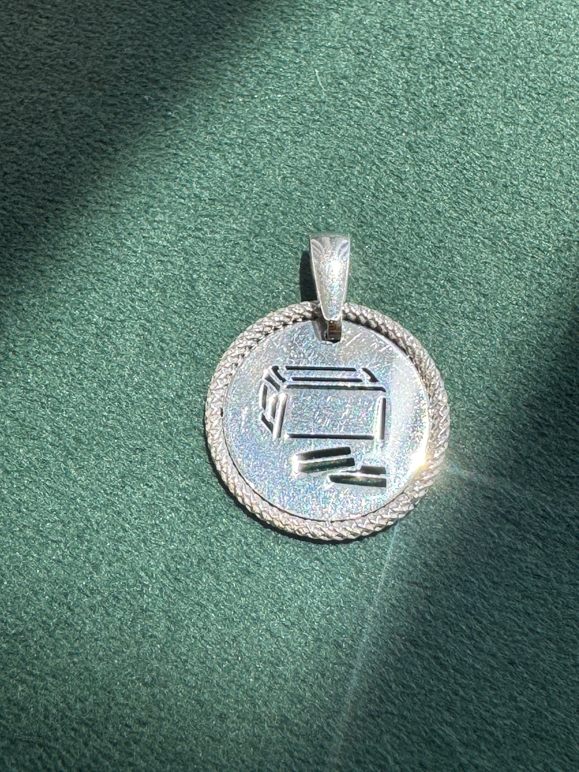 A silver pendant with a circular design, featuring a Hebrew letter 'Shin' engraved in the center and a braided border, resting on a textured green surface.