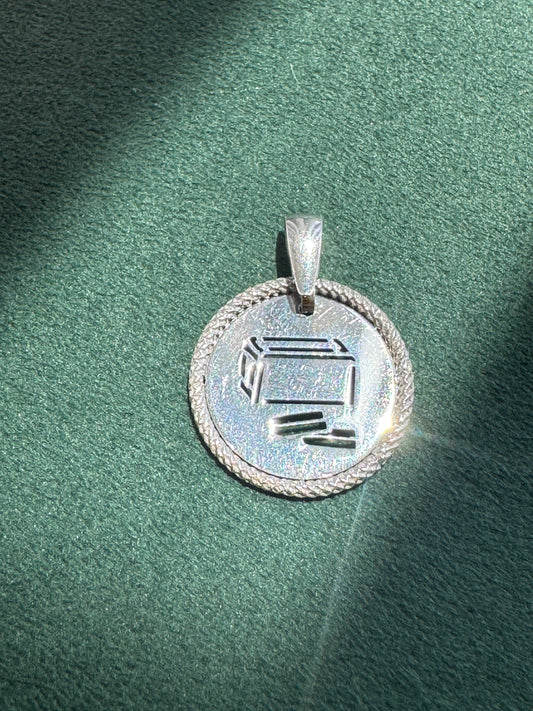A silver pendant with a circular design, featuring a Hebrew letter 'Shin' engraved in the center and a braided border, resting on a textured green surface.