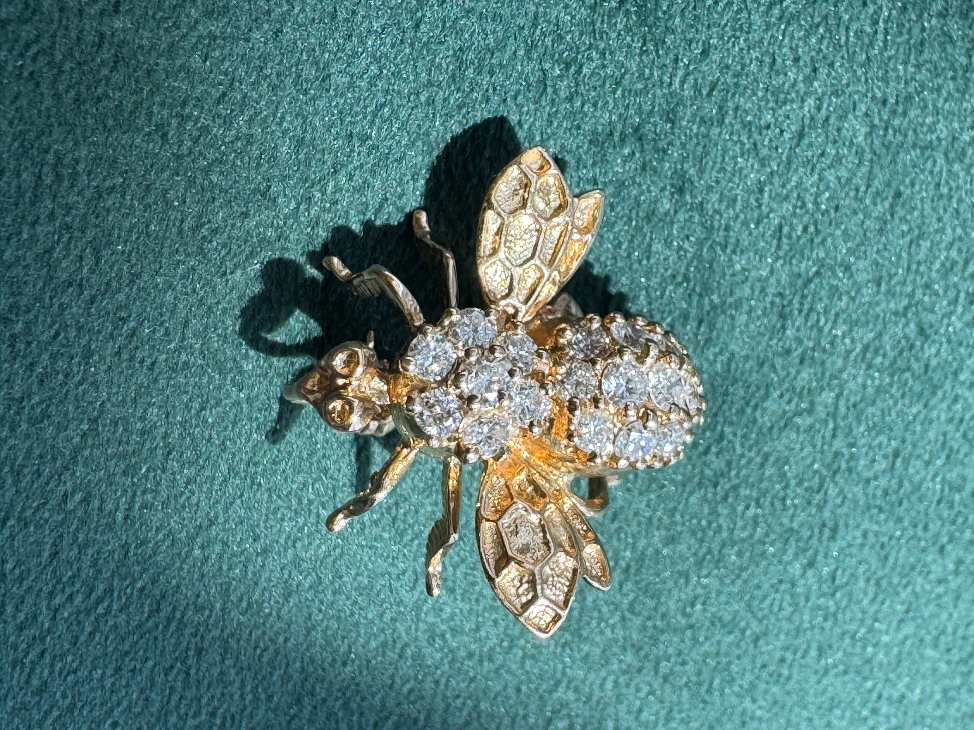 A gold bee-shaped brooch or ring embellished with clear pavé stones on its body and wings, photographed on a textured teal-green background.